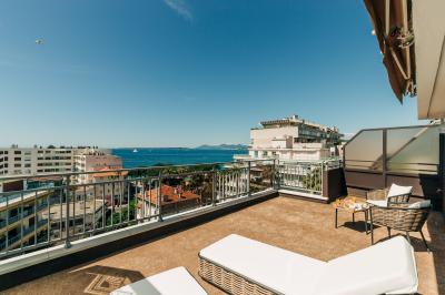 Chambre lumineuse avec lit double, fauteuils, table basse, valise et rideaux blancs laissant entrer la lumière naturelle.