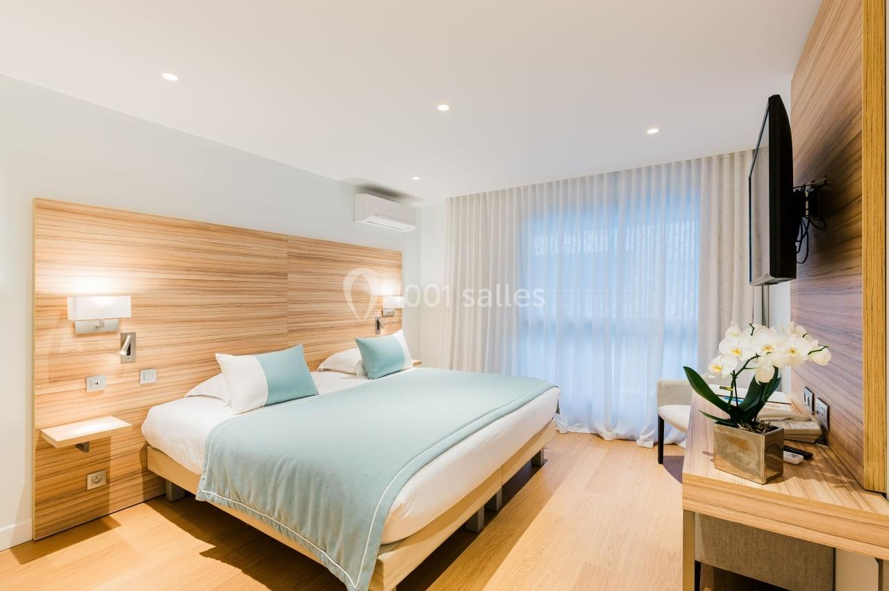 Chambre lumineuse avec deux lits, draps bleu clair, mobilier en bois, télévision murale et orchidée sur une console.