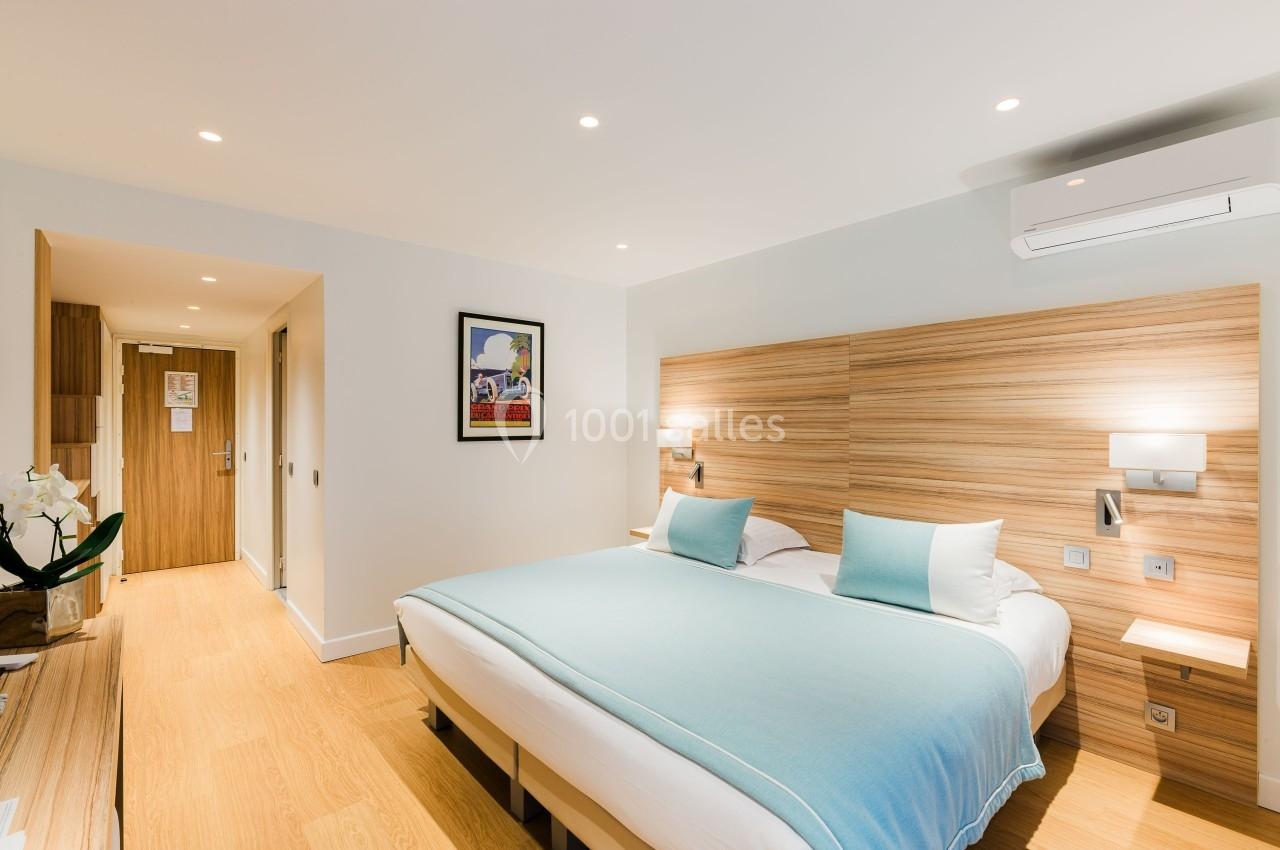 Chambre d'hôtel lumineuse avec lit double, couvre-lit bleu, mobilier en bois clair et éclairage moderne.