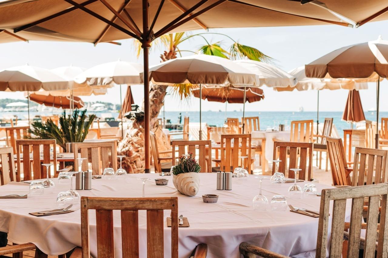 Terrasse en bord de mer avec tables dressées, chaises en bois et parasols beige sous un ciel dégagé.