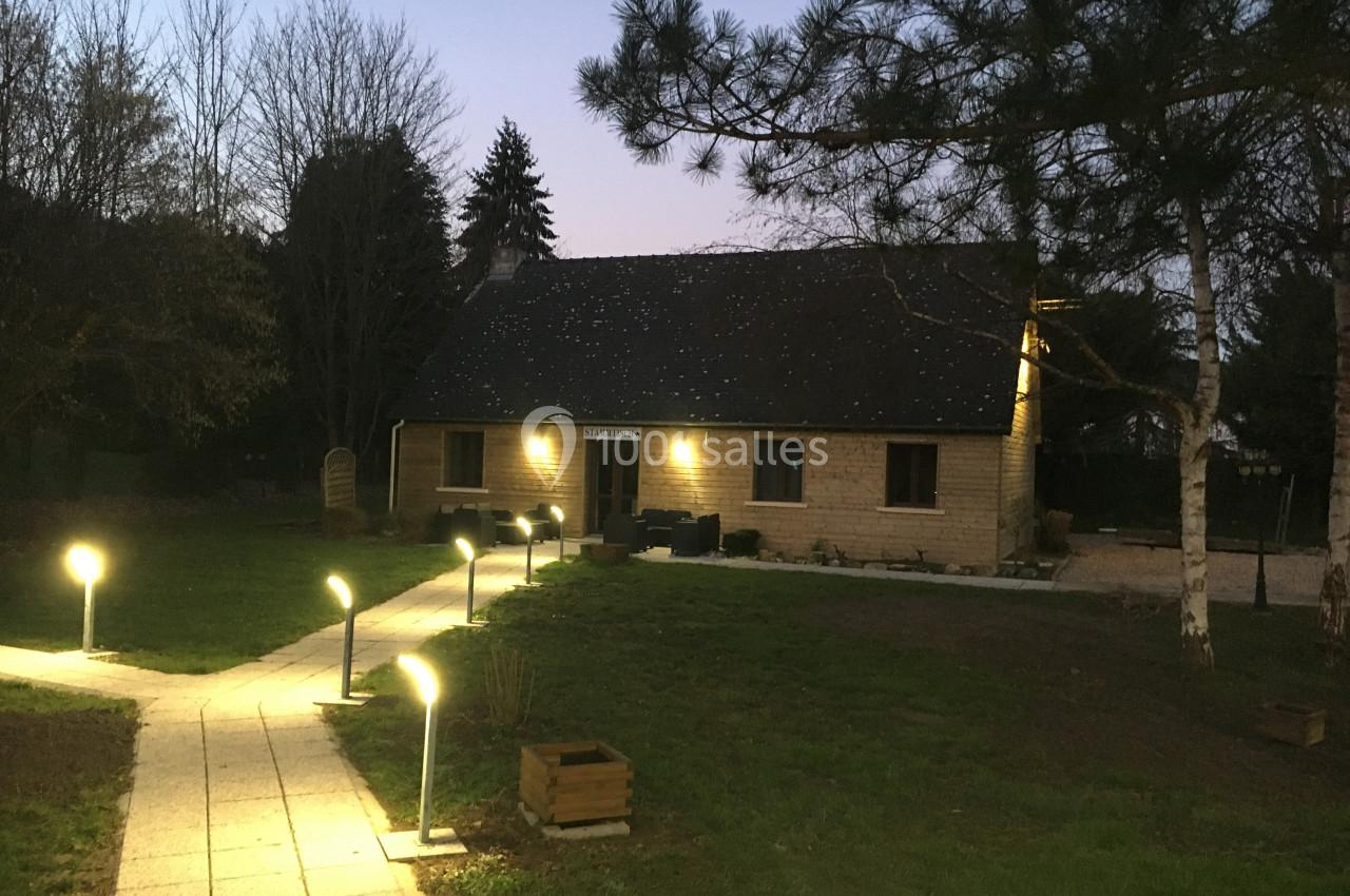 Maison en bois éclairée à la tombée de la nuit, entourée d'arbres et d'un chemin bordé de lampadaires.