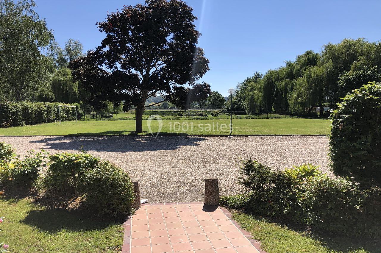 Allée en gravier bordée de verdure menant à un grand jardin avec un arbre central sous un ciel dégagé.