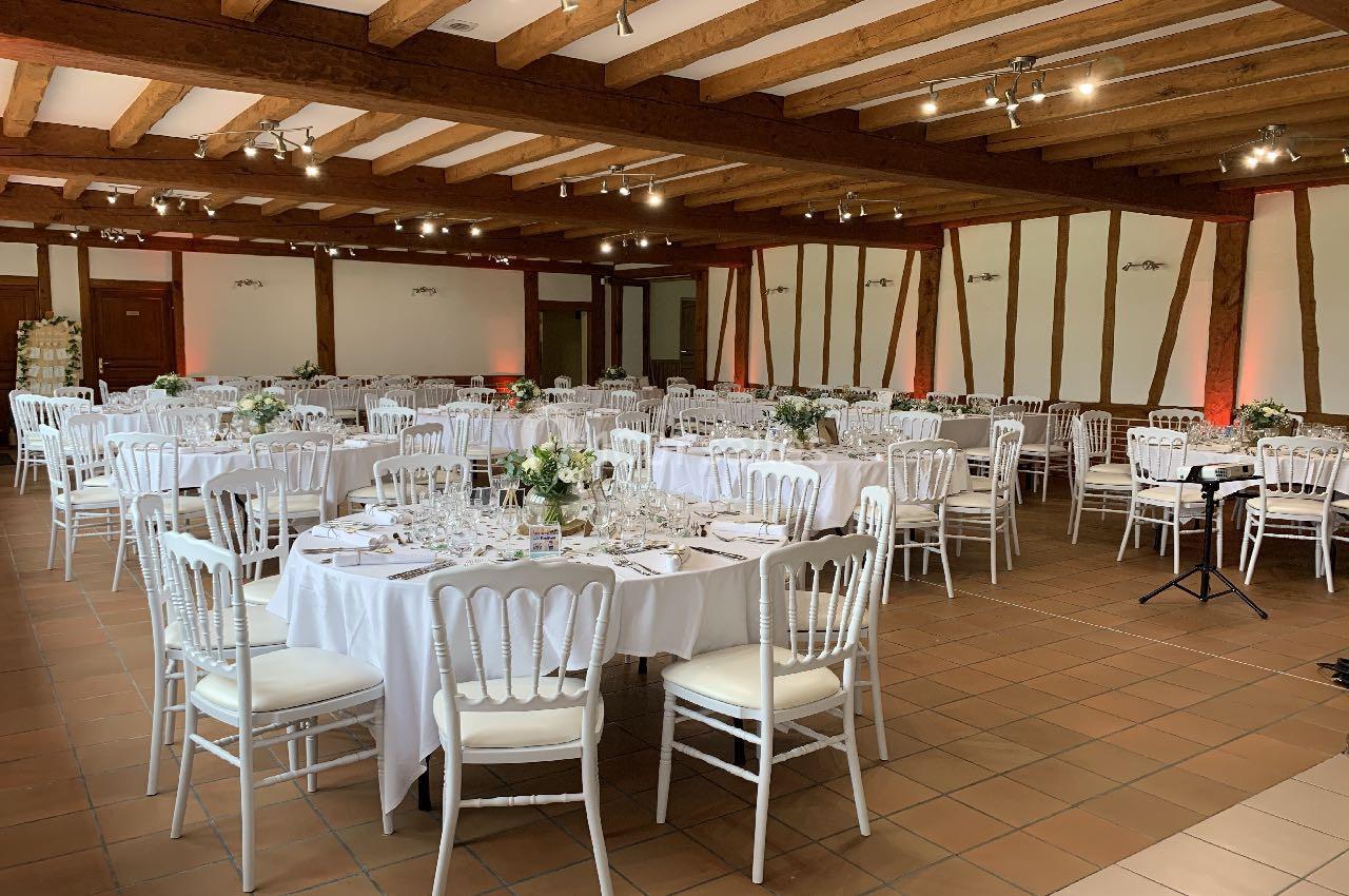 Salle de réception avec tables rondes dressées, nappes blanches, chaises en bois blanc et poutres apparentes au plafond.