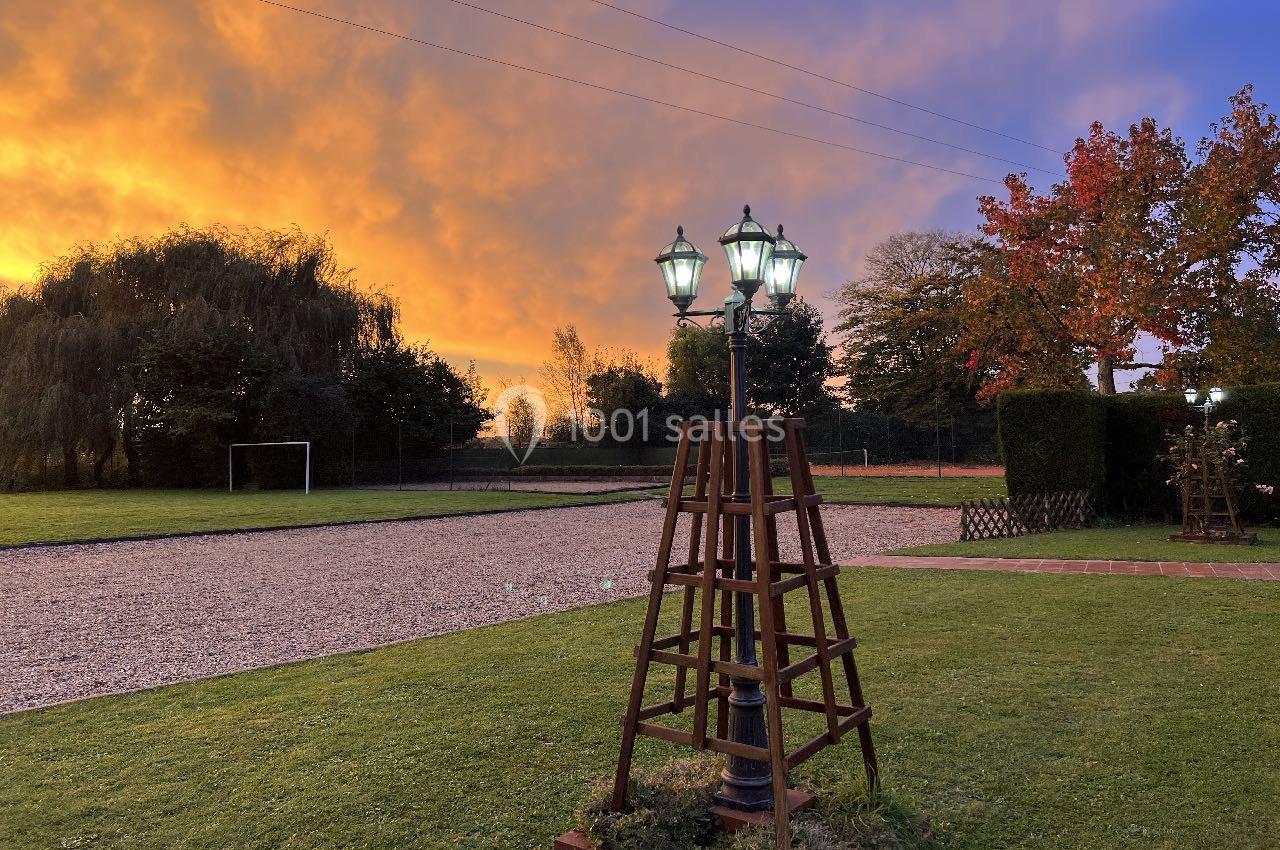 Lampadaire vintage éclairé dans un jardin au coucher du soleil, avec pelouse, arbres et ciel aux teintes orangées.
