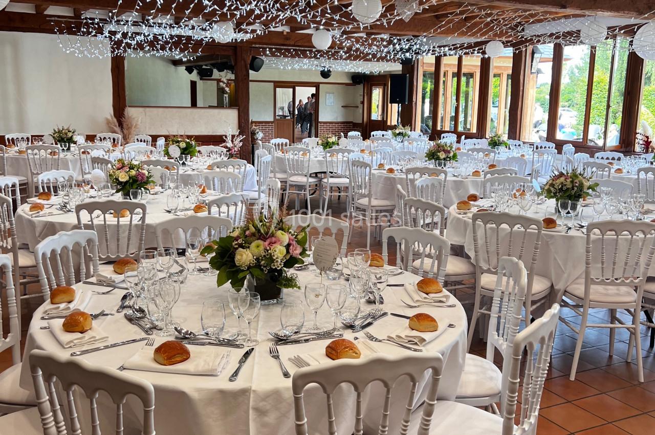 Salle de réception décorée avec des tables rondes dressées, des chaises blanches et des guirlandes lumineuses suspendues.