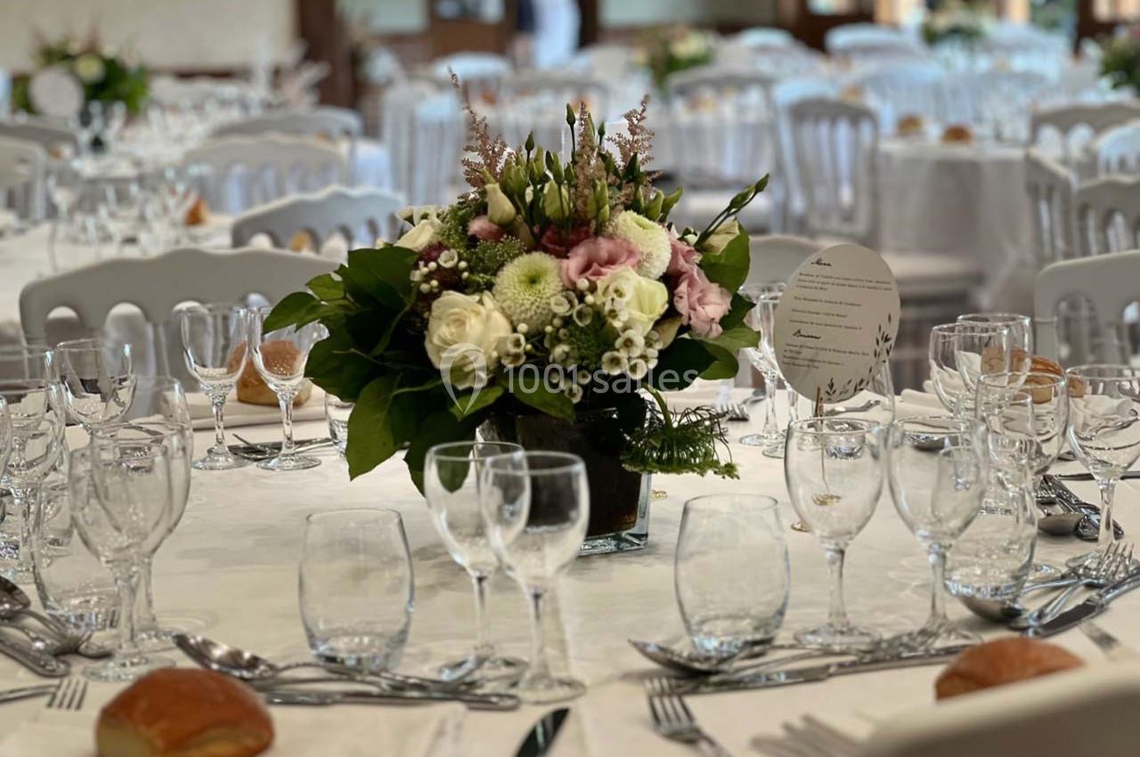 Salle de réception avec tables dressées, centre de table floral, verres, couverts et menus disposés élégamment.
