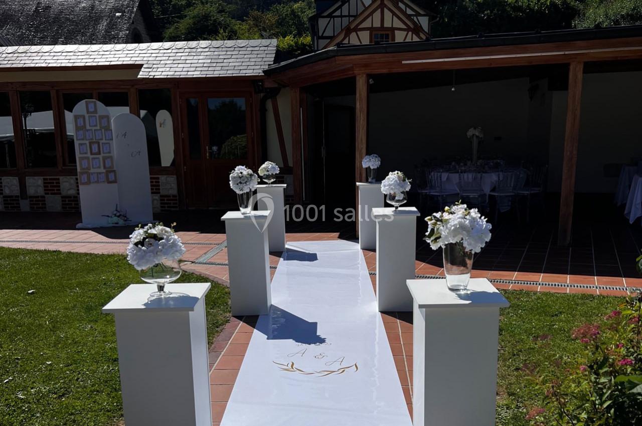 Allée décorée de vases fleuris menant à une salle ouverte, dans un cadre extérieur avec bâtiments en arrière-plan.