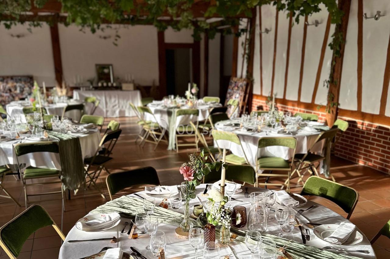 Salle de réception décorée avec des tables rondes dressées, nappes blanches, chaises vertes et feuillage suspendu.