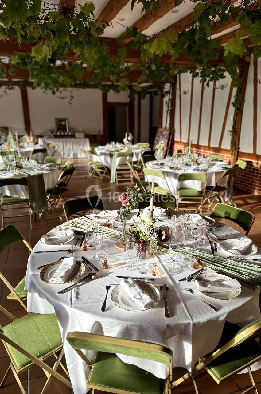 Salle de réception décorée avec des tables rondes dressées, nappes blanches et chaises vertes, sous un plafond orné de…