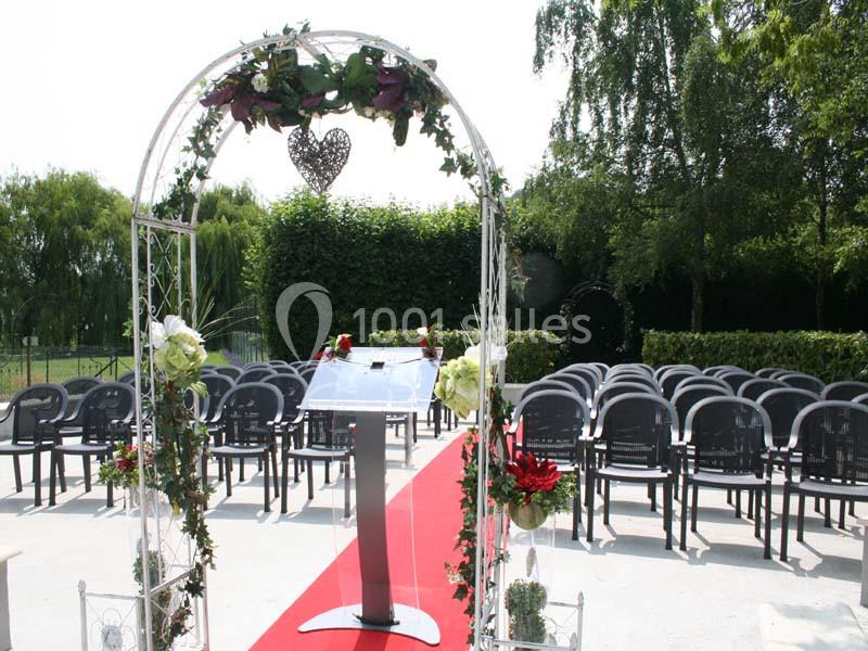 Arche décorée de fleurs et pupitre sur tapis rouge, entourés de chaises noires en extérieur, prêt pour une cérémonie.