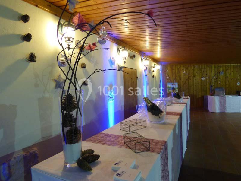 Salle décorée avec des tables alignées, ornées de branches, pommes de pin et bouteilles dans un éclairage tamisé.
