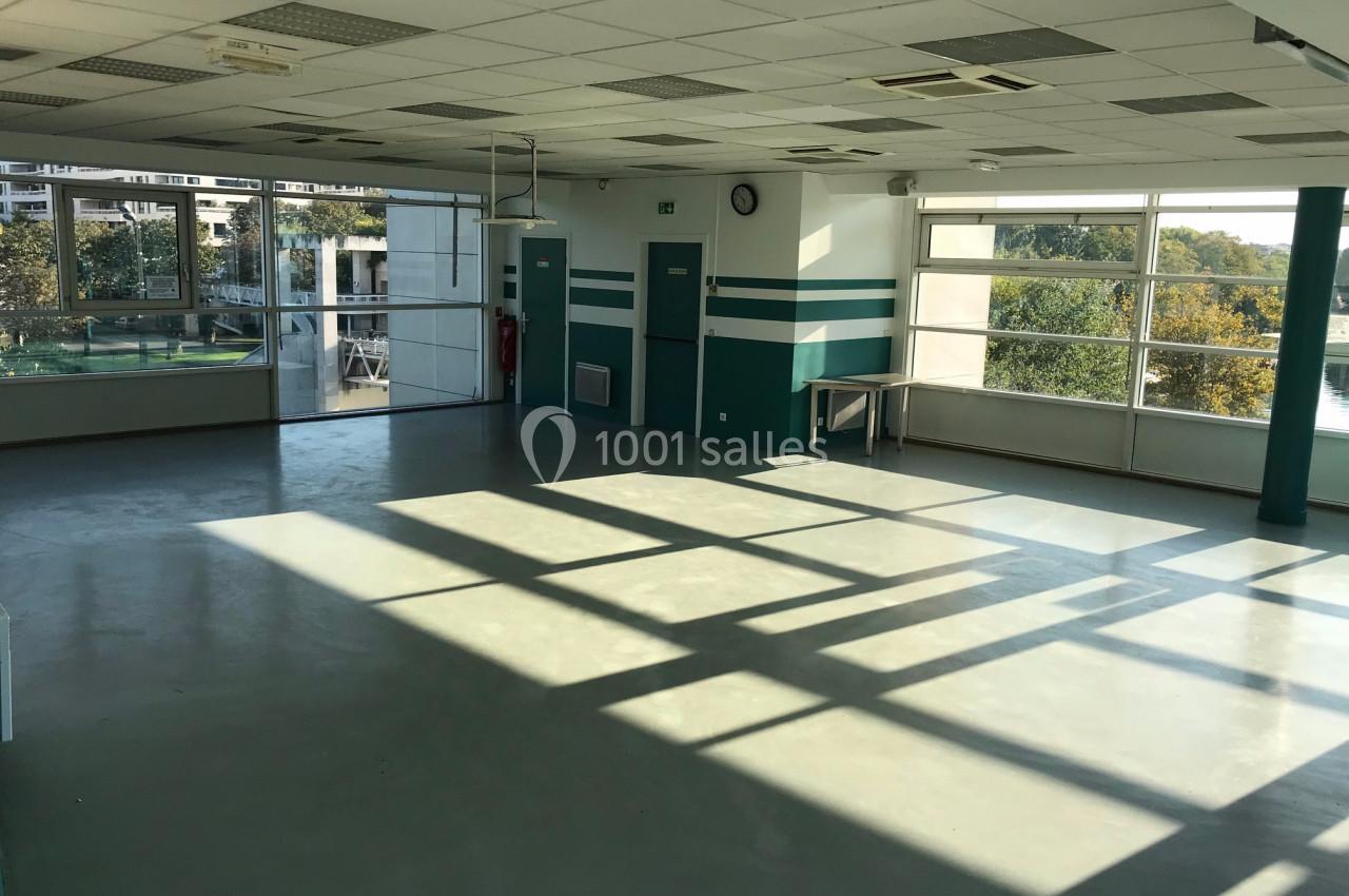 Salle vide avec sol en béton, grandes fenêtres laissant entrer la lumière naturelle et vue sur des arbres à l'extérieur.