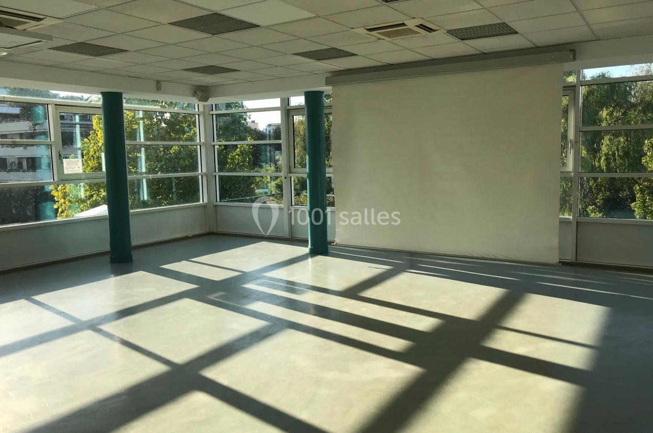 Salle vide lumineuse avec de grandes fenêtres, un sol en béton et un écran blanc fixé au mur.