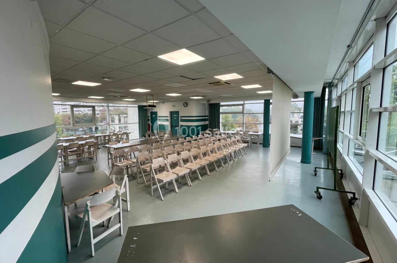 Salle lumineuse avec rangées de chaises blanches, tables et écran de projection, entourée de grandes fenêtres.