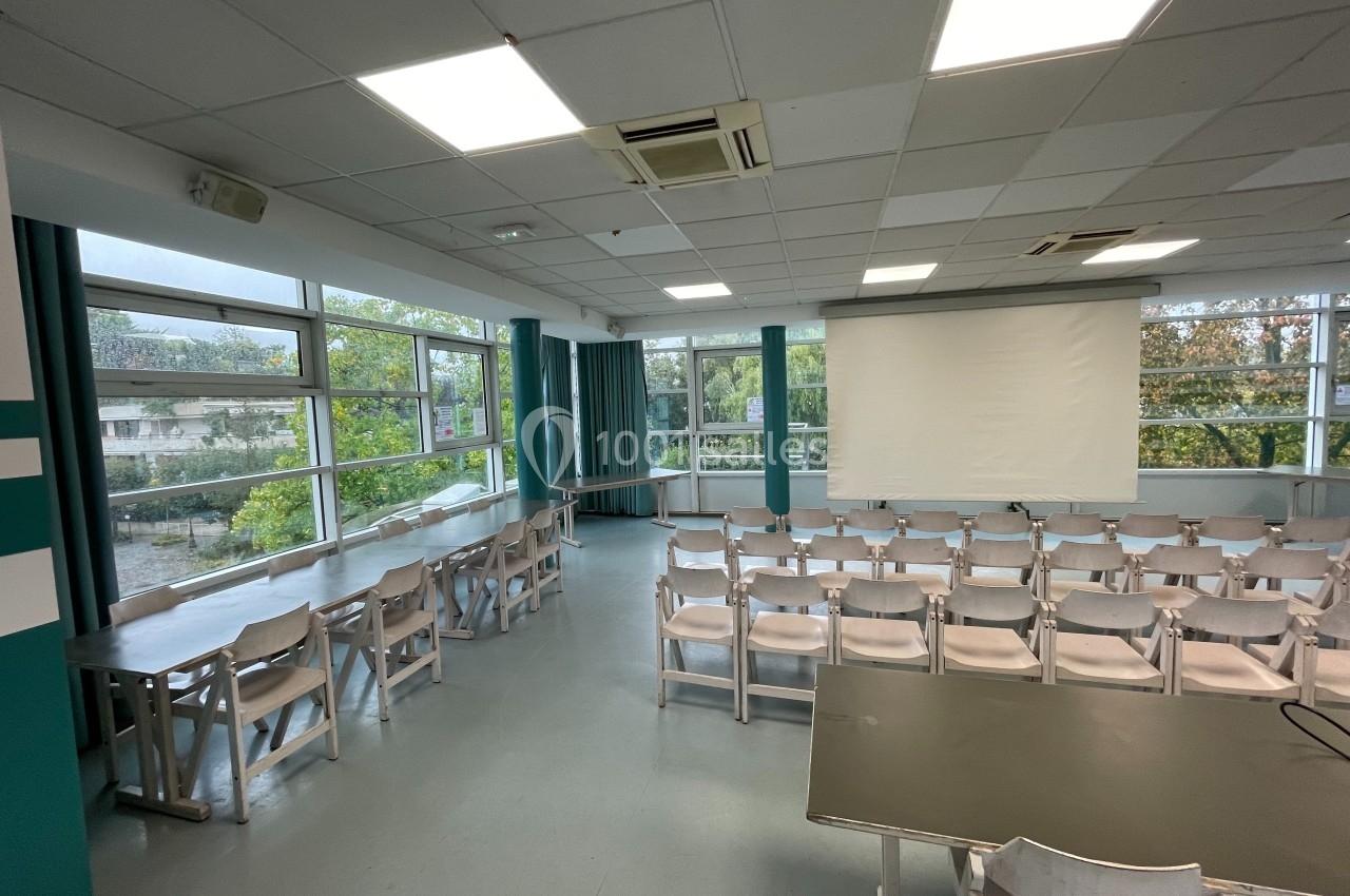 Salle de réunion lumineuse avec des rangées de chaises, des tables et un écran de projection devant des fenêtres donnant sur…