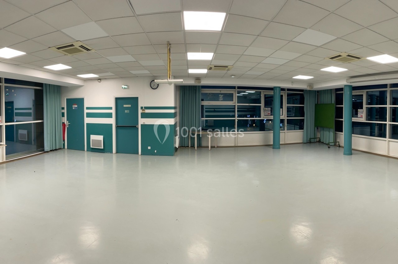 Salle vide avec sol clair, murs blancs et verts, plafond à dalles blanches et grandes fenêtres donnant sur un espace sombre.