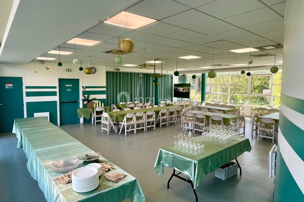 Salle décorée pour un événement, avec tables dressées, nappes vertes, chaises blanches et lanternes suspendues.
