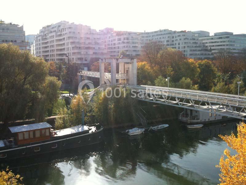 Passerelle piétonne traversant une rivière bordée d'arbres, avec des immeubles en arrière-plan.