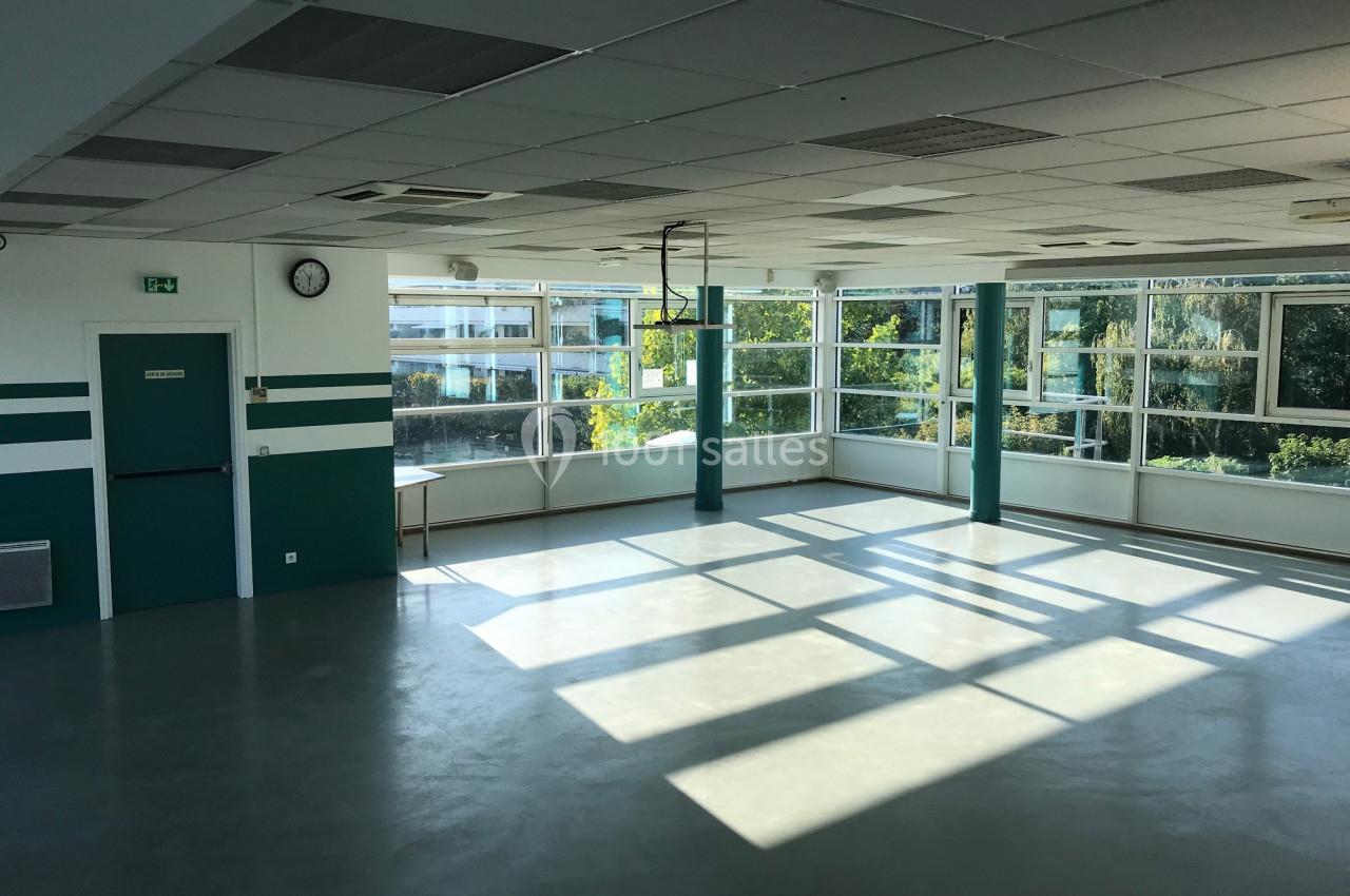 Salle lumineuse avec de grandes fenêtres, murs blancs et verts, sol clair, et vue sur des arbres à l'extérieur.