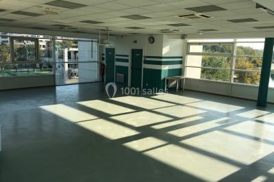 Salle vide avec sol clair, murs verts et blancs, grandes fenêtres, éclairage au plafond et mobilier minimal.