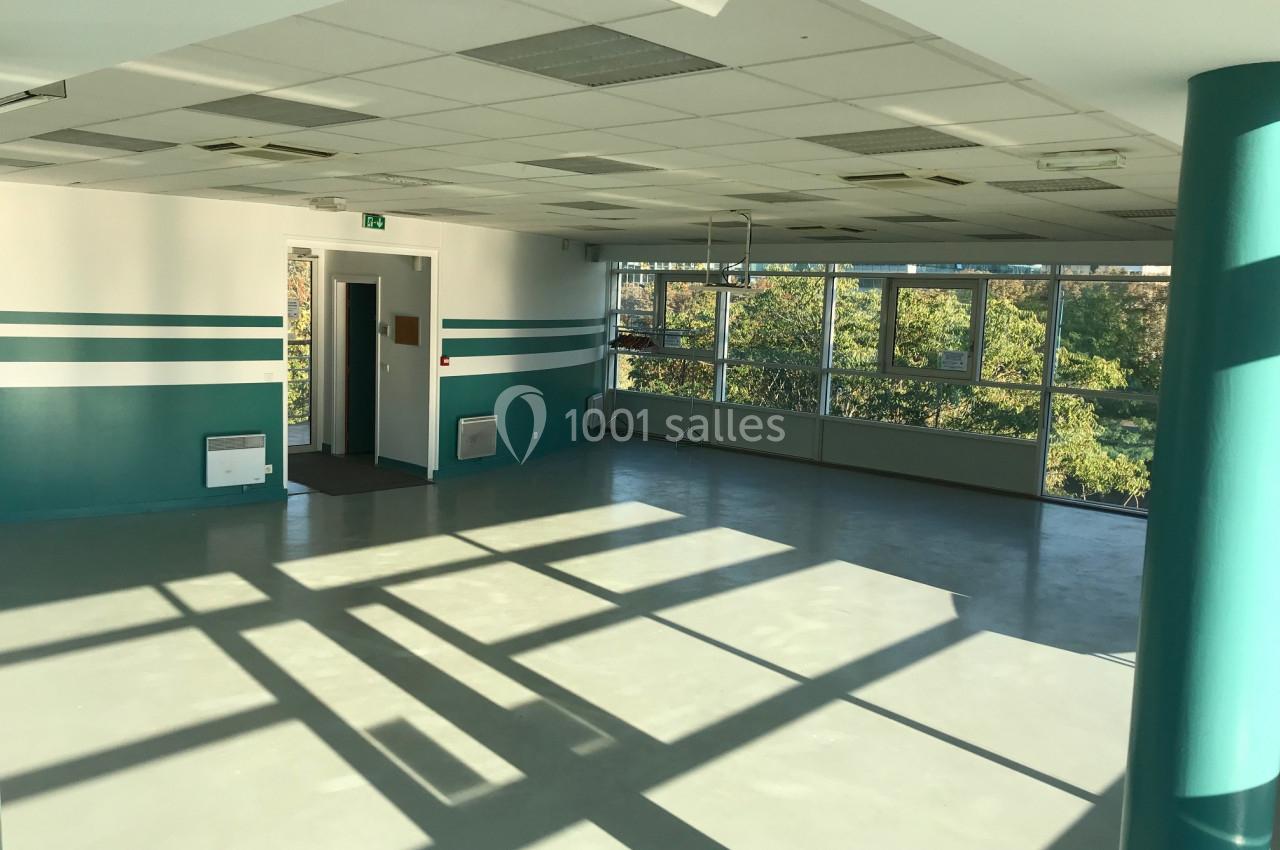 Salle vide avec sol en béton, murs blancs et verts, grandes fenêtres laissant entrer la lumière naturelle.