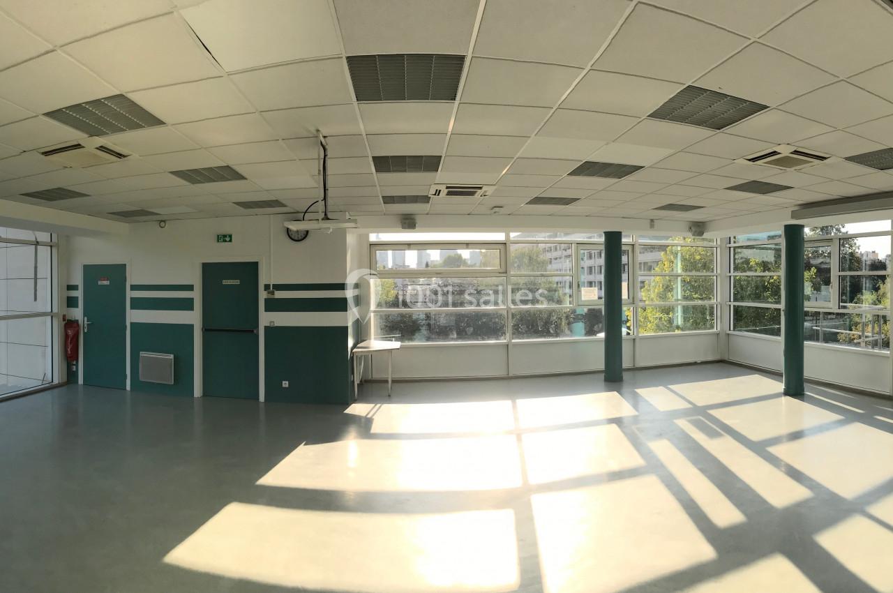 Salle lumineuse avec de grandes baies vitrées, vue sur des arbres et des bâtiments, sol clair et plafond à dalles.