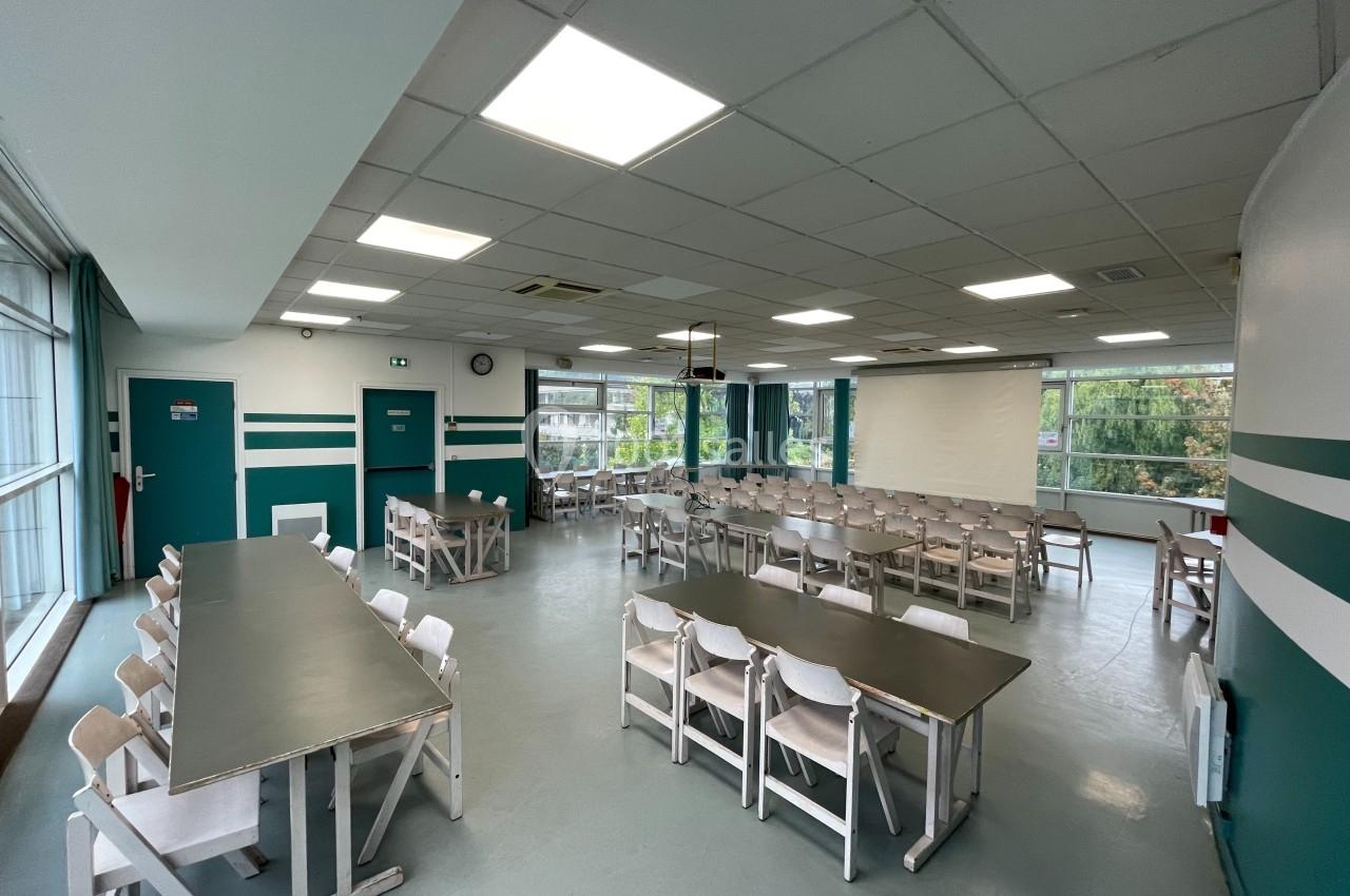 Salle de restauration lumineuse avec tables et chaises alignées, grandes fenêtres donnant sur un espace arboré.