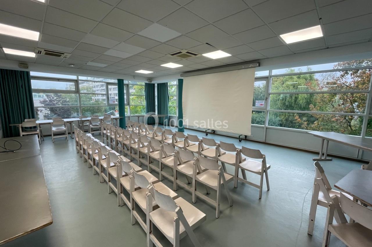Salle de conférence lumineuse avec des rangées de chaises blanches, un écran de projection et de grandes fenêtres donnant…
