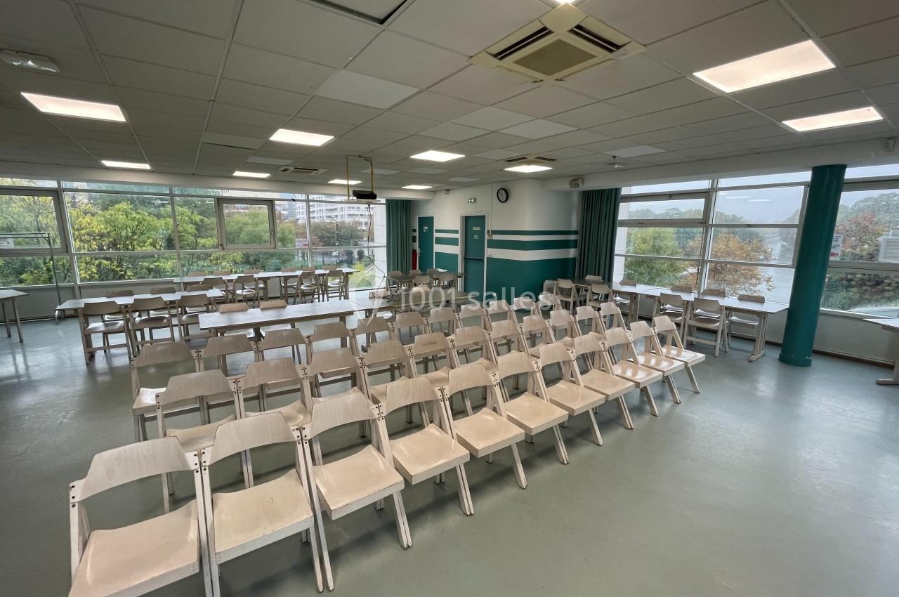 Salle lumineuse avec rangées de chaises blanches alignées, grandes fenêtres donnant sur un paysage arboré.
