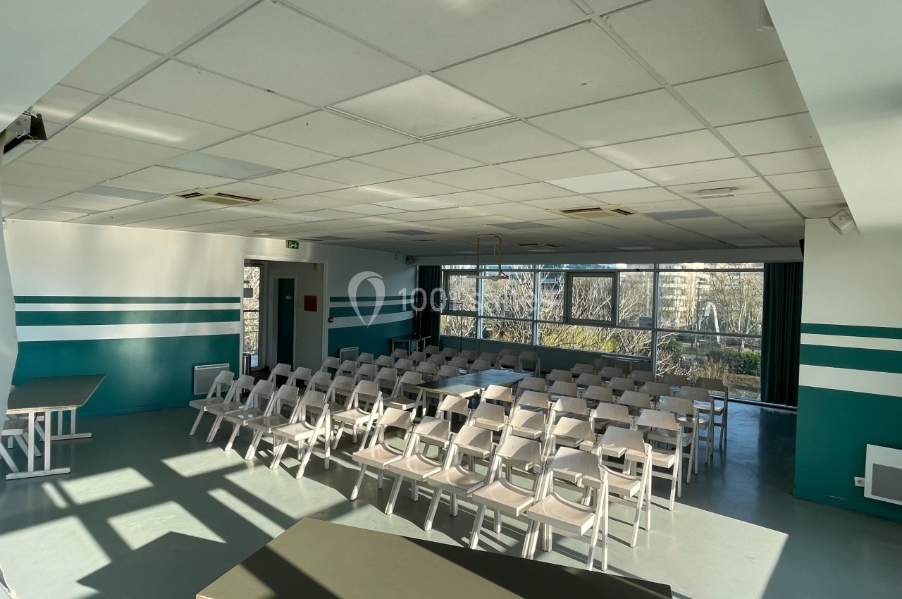 Salle lumineuse avec des rangées de chaises blanches alignées, grandes fenêtres donnant sur un paysage extérieur.