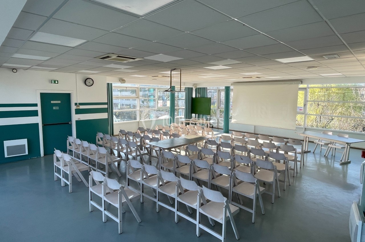 Salle lumineuse avec des rangées de chaises blanches disposées face à un écran de projection et des fenêtres.