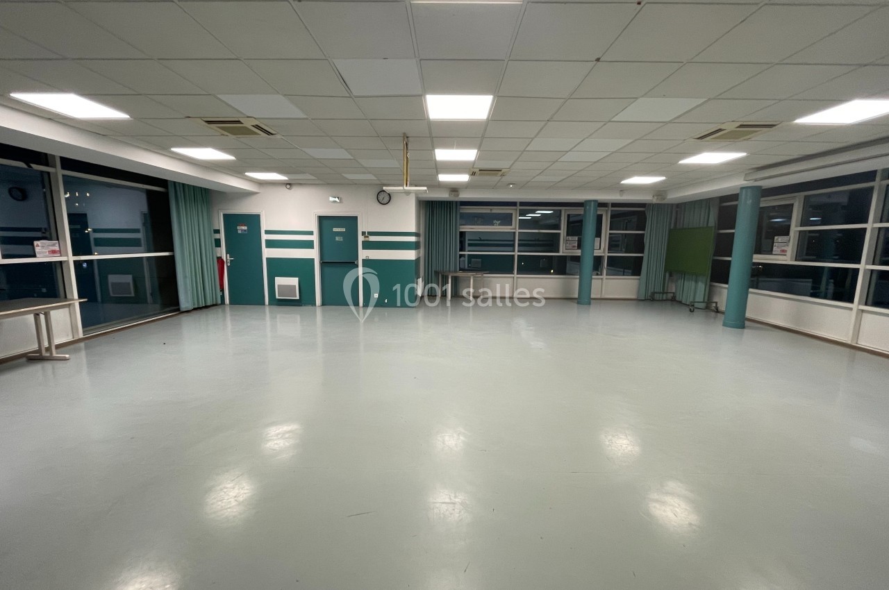 Salle vide avec sol clair, murs verts et blancs, grandes fenêtres, éclairage au plafond et mobilier minimal.