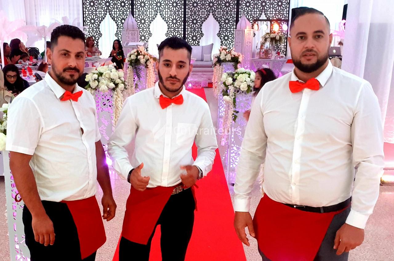 Trois serveurs en chemise blanche et nœud papillon rouge posent dans une salle décorée pour un événement festif.