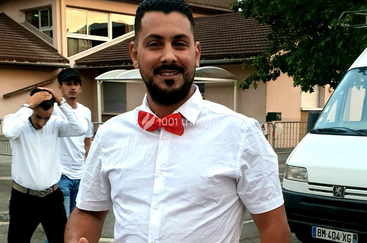 Homme souriant portant une chemise blanche et un nœud papillon rouge, debout à l'extérieur près de bâtiments et d'une…