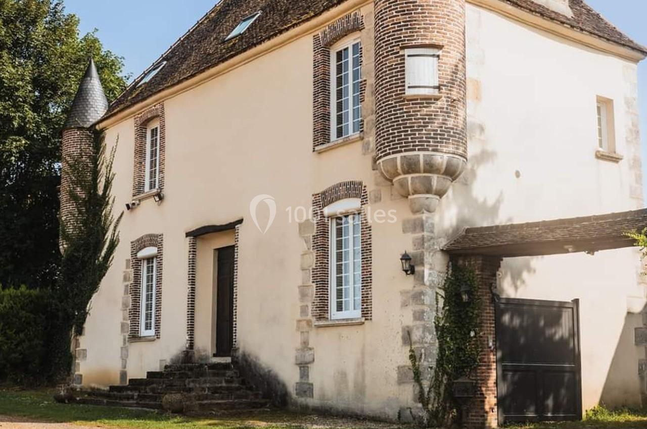 Maison ancienne en pierre et briques avec une petite tour, entourée de verdure, sous un ciel dégagé.