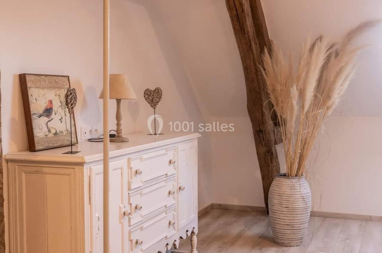 Commode blanche avec lampe et décorations, vase en céramique avec herbes séchées, poutre en bois visible.