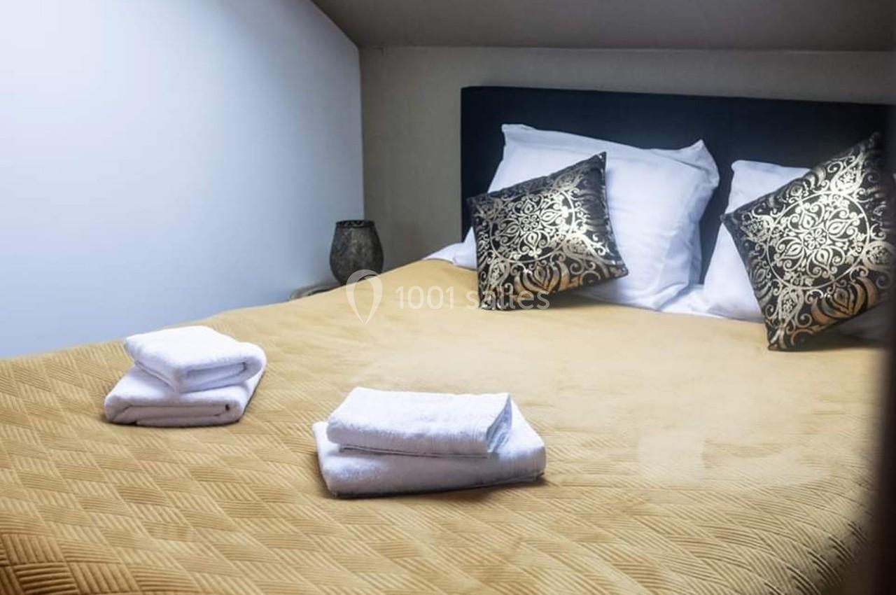 Lit double avec couvre-lit beige, deux oreillers décoratifs noirs et dorés, et serviettes blanches pliées au premier plan.