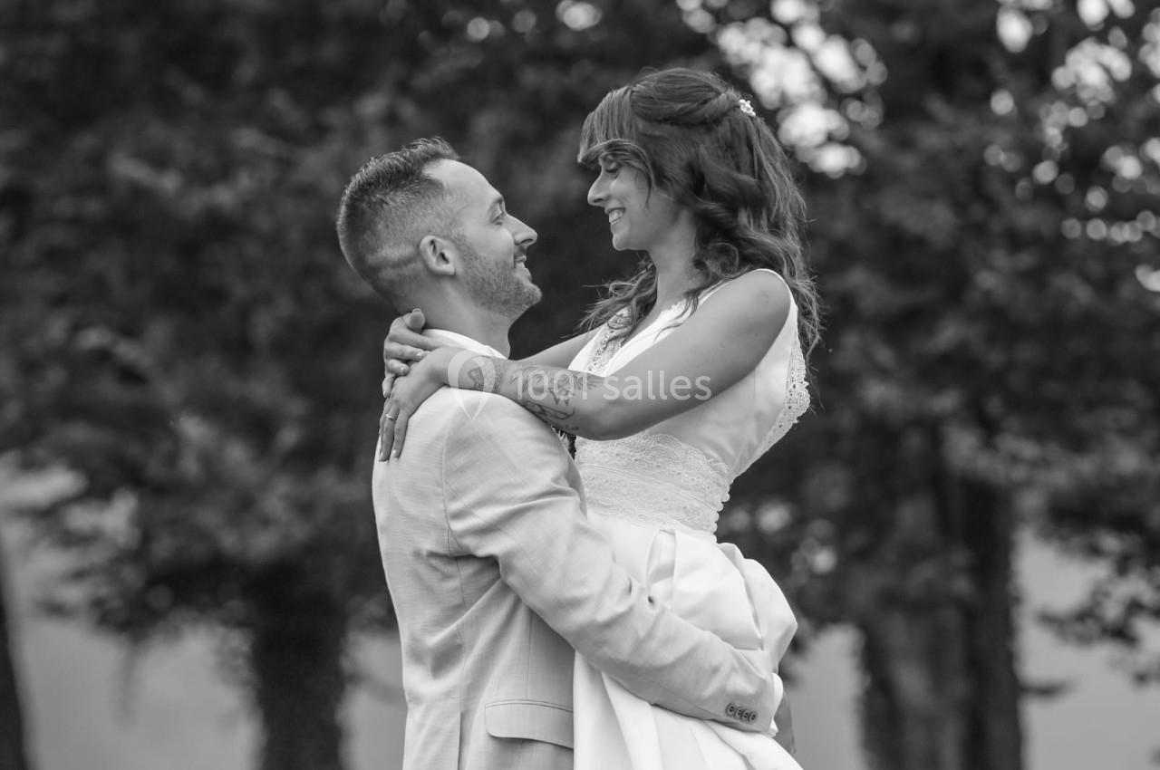 Un homme en costume porte une femme en robe de mariée, tous deux souriants, dans un cadre extérieur arboré.