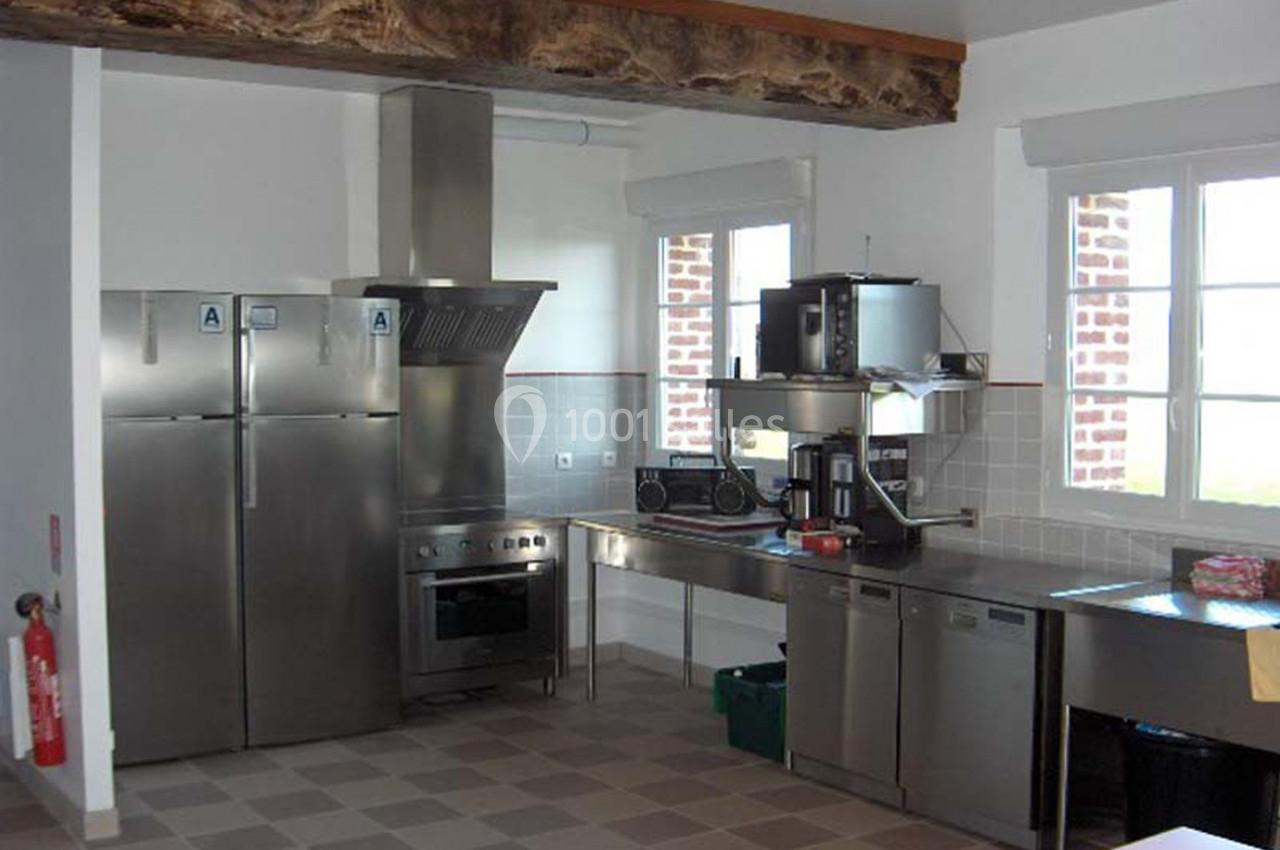 Cuisine équipée avec électroménagers en inox, plan de travail en métal, carrelage au sol et poutre apparente au plafond.