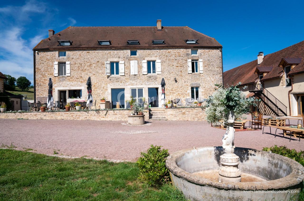 Façade d'une grande maison en pierre avec terrasse, fontaine au premier plan et ciel bleu en arrière-plan.