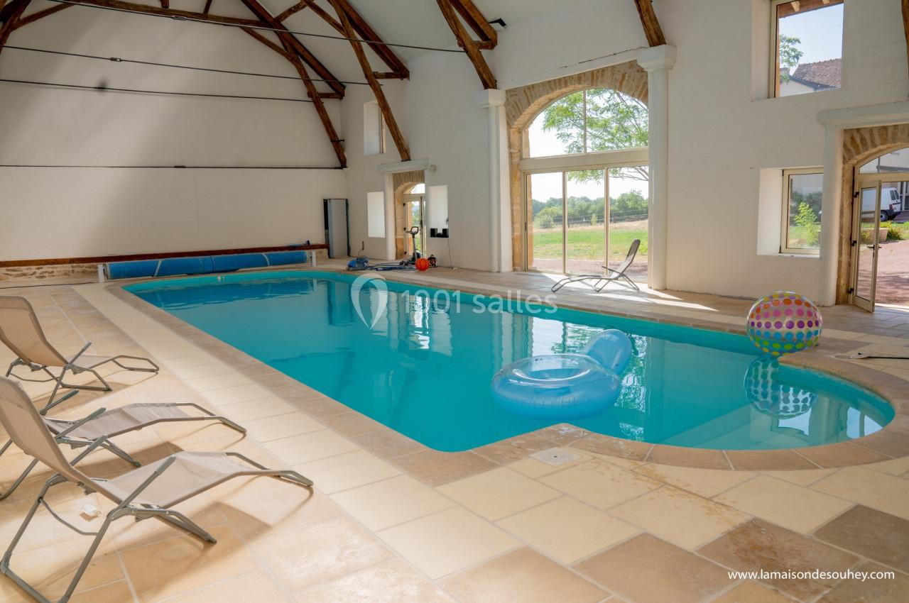 Piscine intérieure lumineuse avec transats, jouets flottants et grandes baies vitrées donnant sur un jardin.