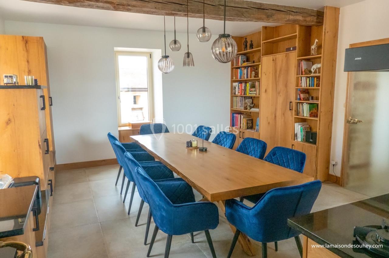 Salle à manger lumineuse avec une grande table en bois, chaises bleues, étagères remplies de livres et suspensions modernes.