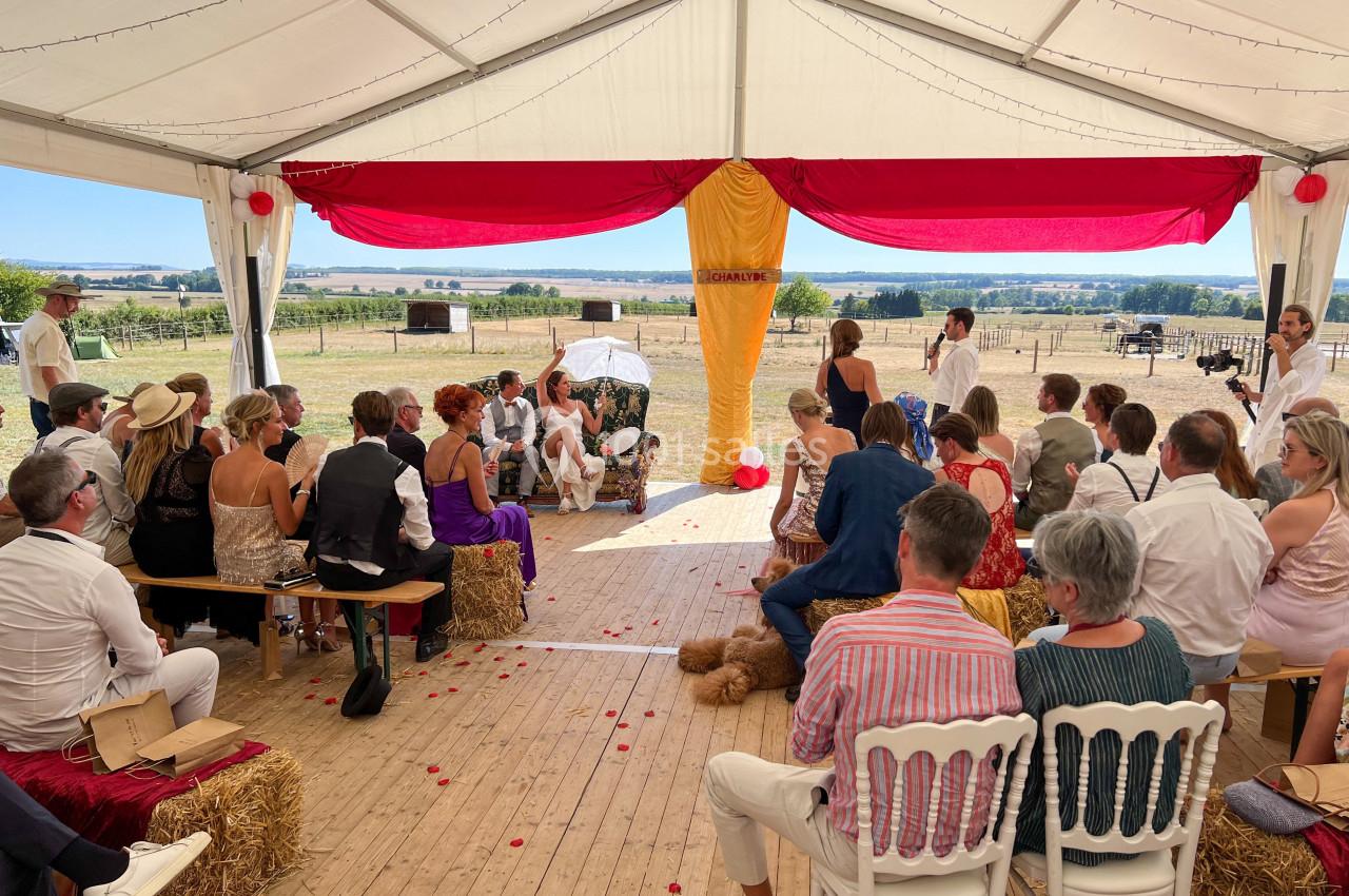 Cérémonie en plein air sous une tente décorée, avec des invités assis sur des bottes de paille et des chaises.