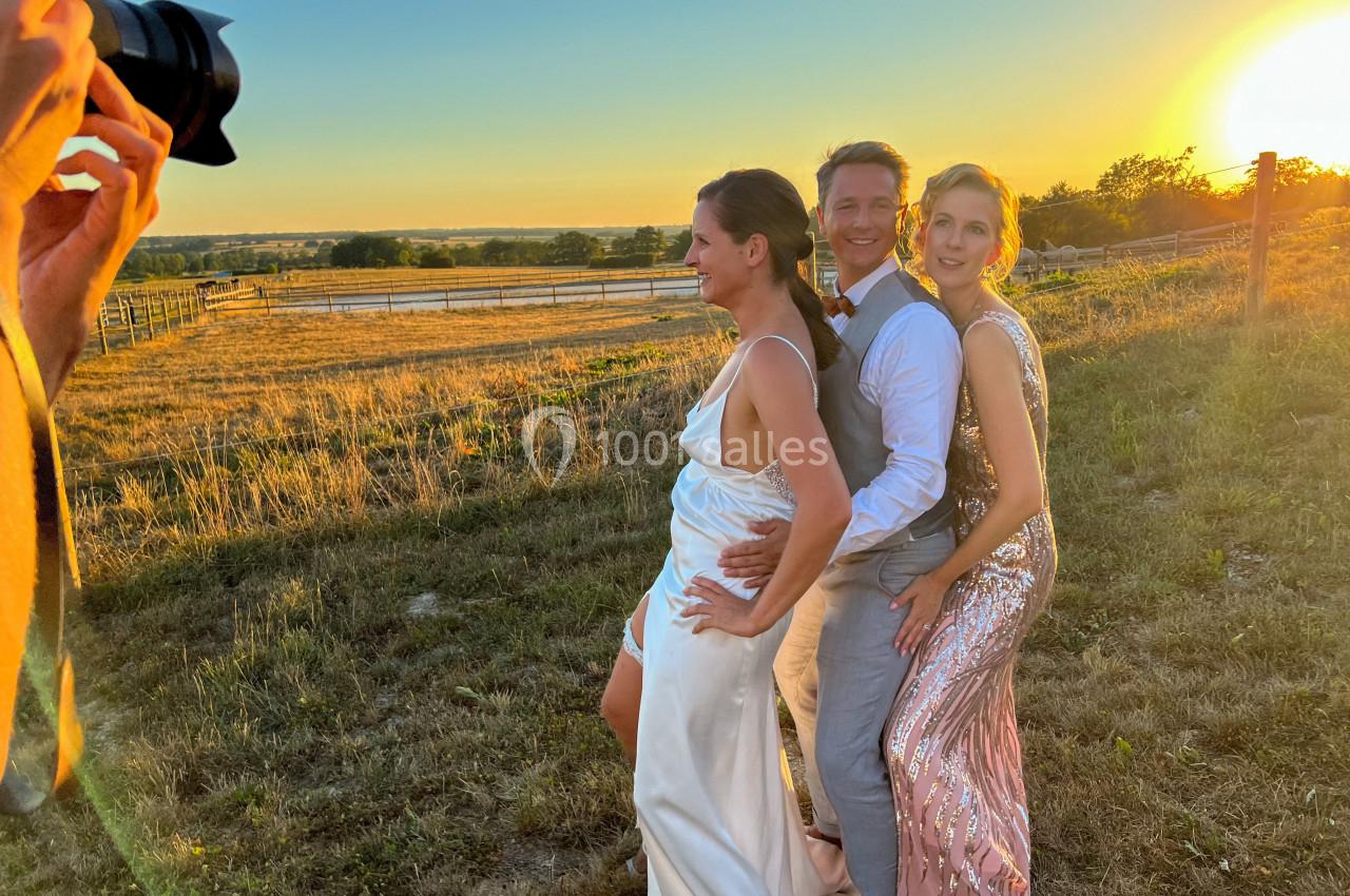 Trois personnes posent en tenue élégante dans un champ au coucher du soleil, avec un photographe au premier plan.