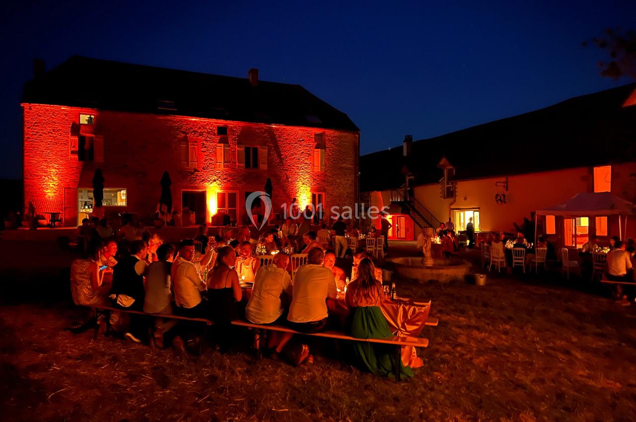 Groupe de personnes dînant en plein air devant un bâtiment en pierre éclairé par des lumières rouges, de nuit.