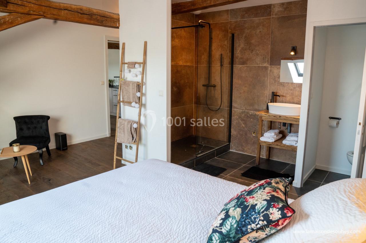 Chambre lumineuse avec lit double, coin salon, salle de bain ouverte avec douche à l'italienne et lavabo sur meuble en bois.