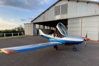 Un homme assis dans un hélicoptère au sol, souriant, avec un coucher de soleil et un champ en arrière-plan.