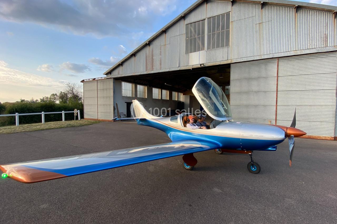 Avion léger biplace au sol devant un hangar métallique, cockpit ouvert avec deux personnes à bord.