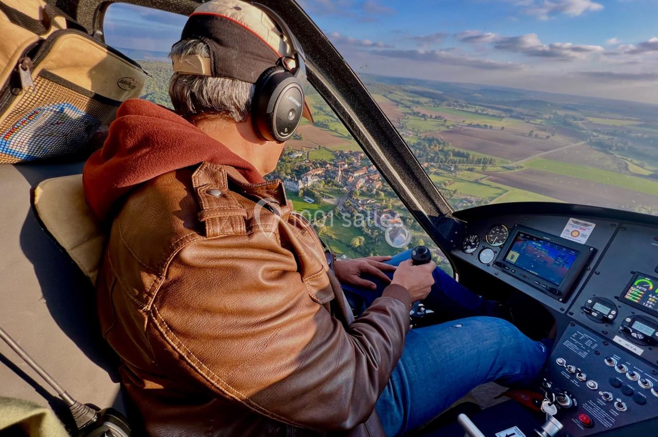 Un pilote en veste marron manœuvre un hélicoptère au-dessus d'une campagne verdoyante par temps clair.