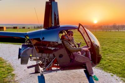 Un homme assis dans un hélicoptère au sol, souriant, avec un coucher de soleil et un champ en arrière-plan.
